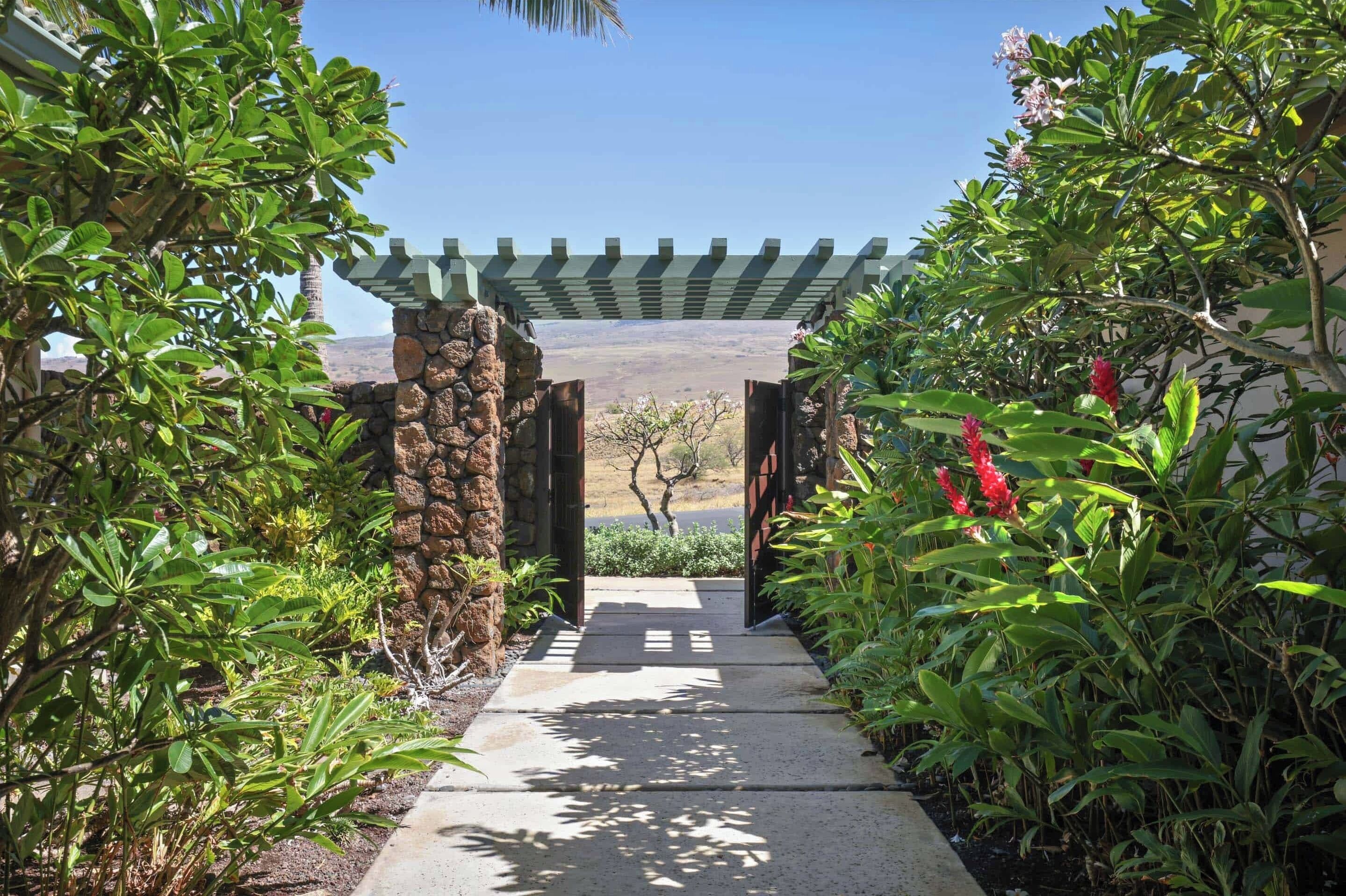 A pathway leading through a garden with a pergola overhead, surrounded by lush greenery and a stone wall.