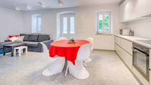Ein modernes Wohnzimmer mit Essbereich, einem grauen Sofa, einer roten Tischdecke und einer Küche mit weißen Schränken und Geräten.