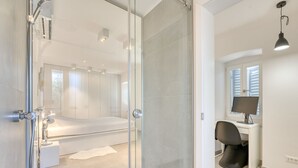 Ein modernes Schlafzimmer mit Duschkabine aus Glas, einem Bett, einem Schreibtisch mit Computer und einem Fenster mit Jalousien.