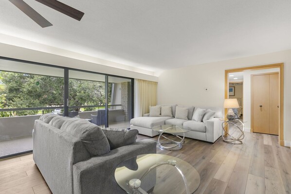 Una sala de estar moderna con un sofá gris, un sofá blanco y una mesa de centro de vidrio. Hay un balcón con puerta de vidrio y vistas a árboles.