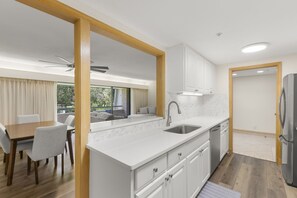 Una cocina moderna con armarios blancos, un lavamanos de acero inoxidable y un horno empotrado. Cuenta con una zona de comedor con mesa y sillas, y vistas a zonas verdes al exterior.