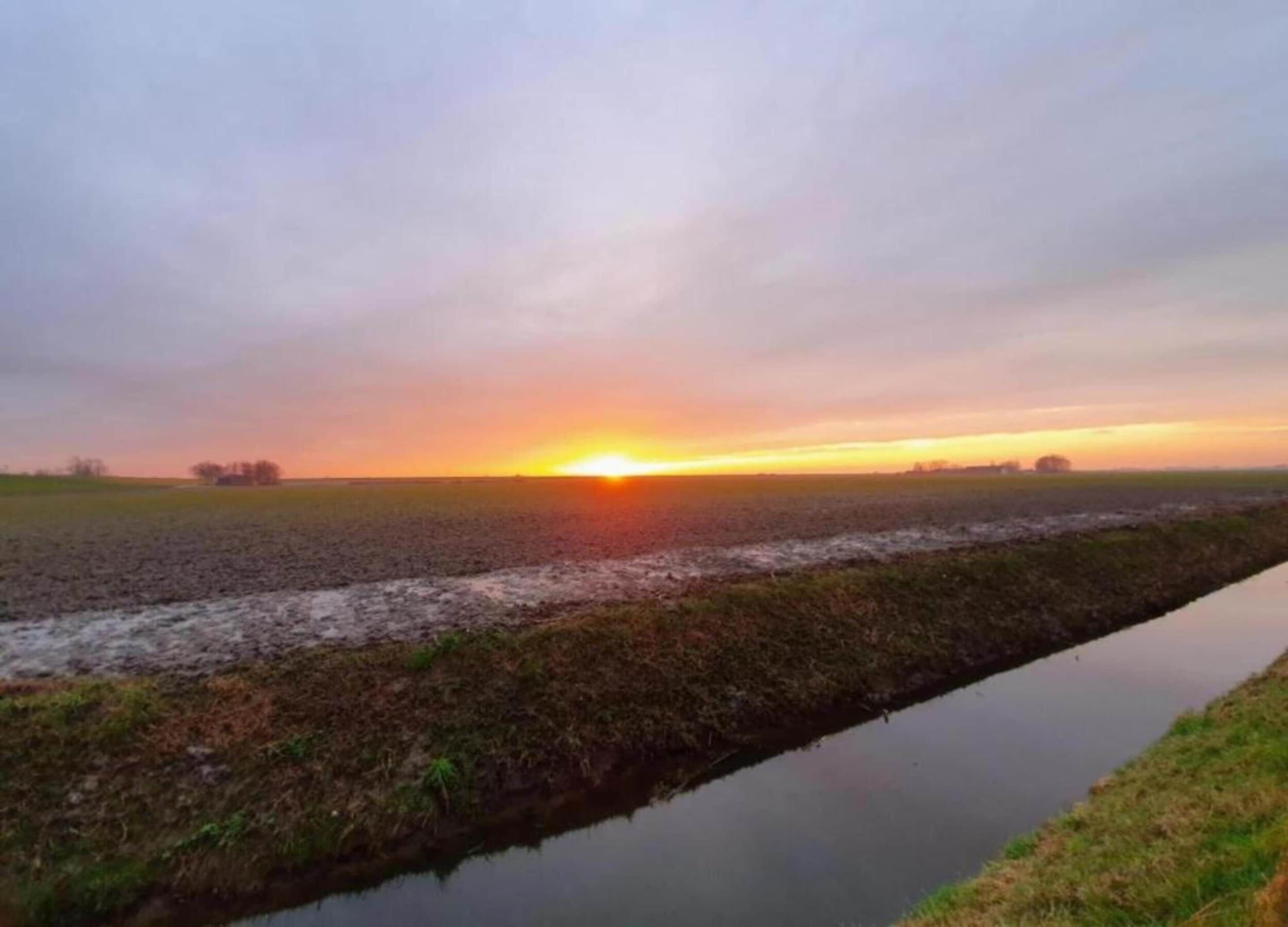 Ein Sonnenuntergang über einer ebenen Landschaft mit einem schmalen Wasserlauf.