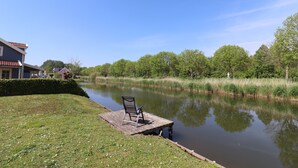 A calm river with a dock and a chair, surrounded by greenery and a house. - Bungalow in Zuytland Buiten by the Water (Simonshaven)