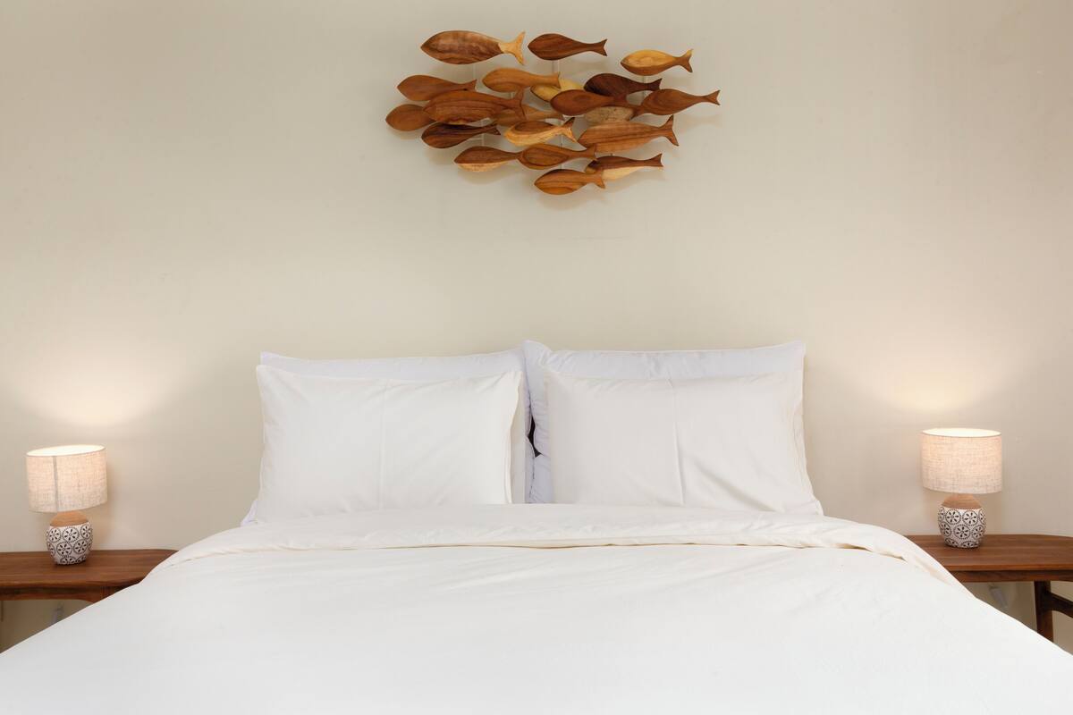 Una cama con ropa blanca, dos mesitas de noche con lámparas y un cuadro de peces.
