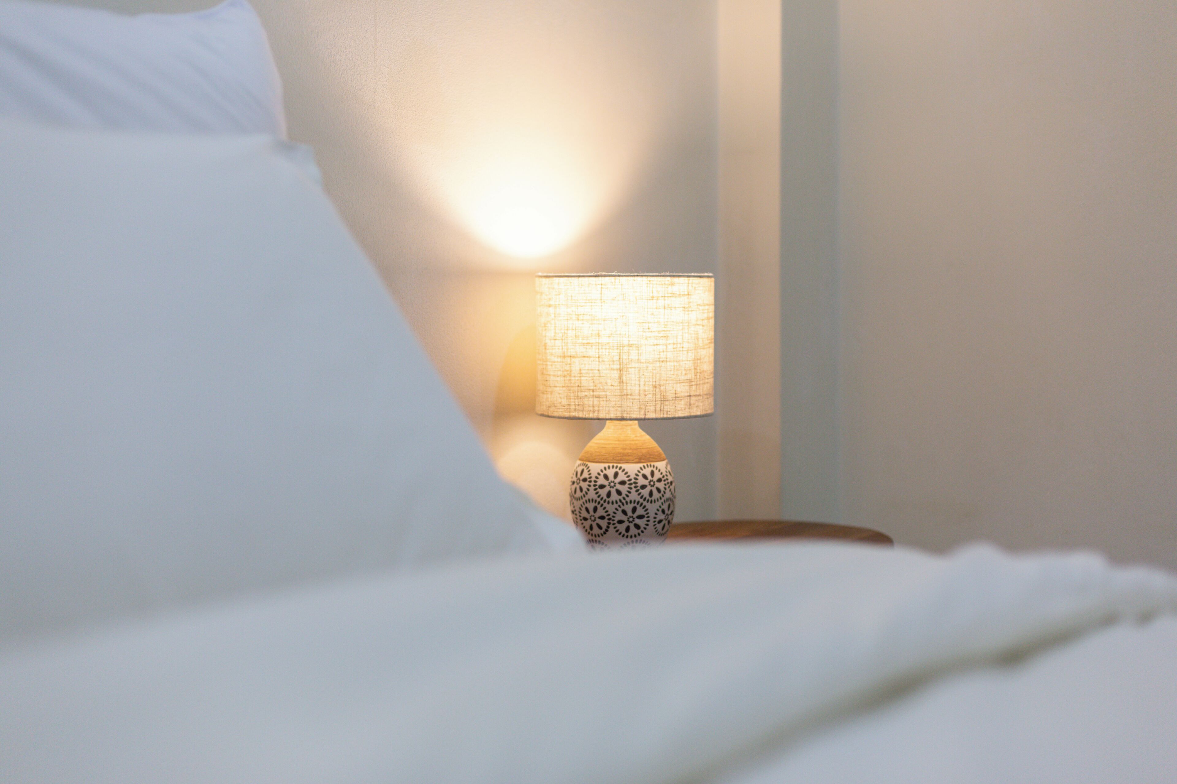 A neatly made bed with white linens, a bedside lamp with a patterned shade, and a wall with a soft glow.