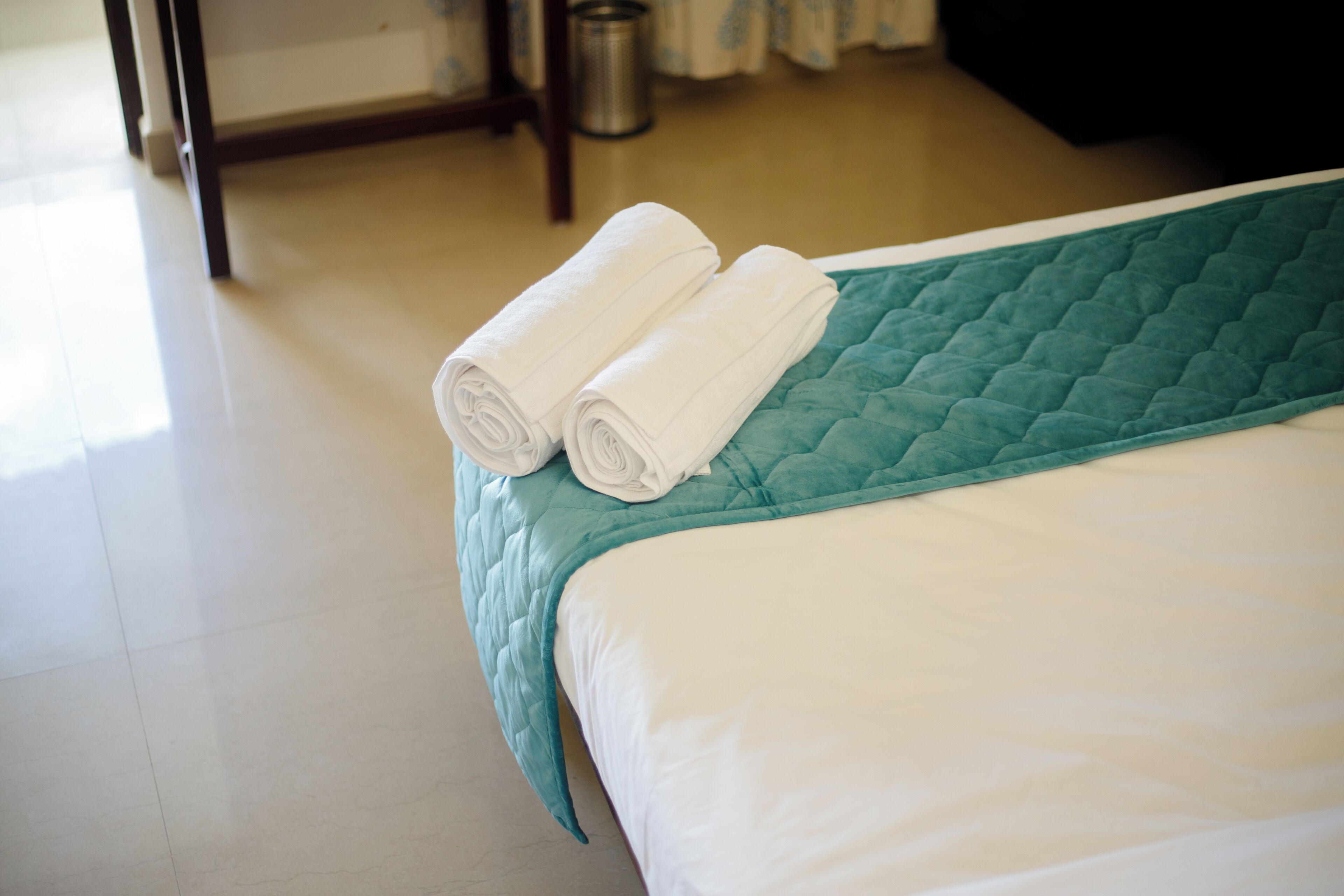 A neatly made bed with a teal quilt and two rolled white towels on the headboard.