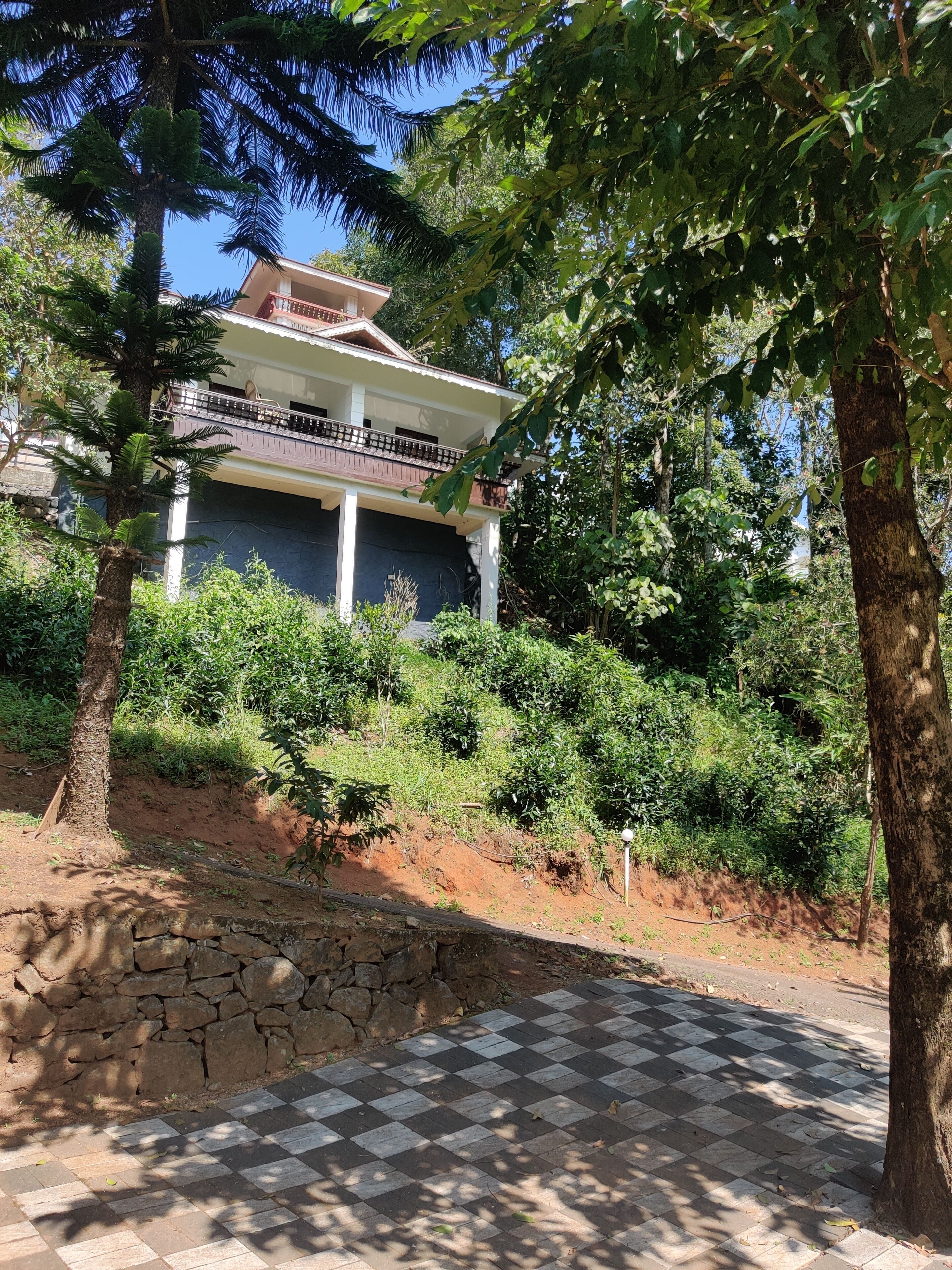 Une maison dotée d’un balcon, entourée d’une végétation luxuriante et d’un mur de pierre.