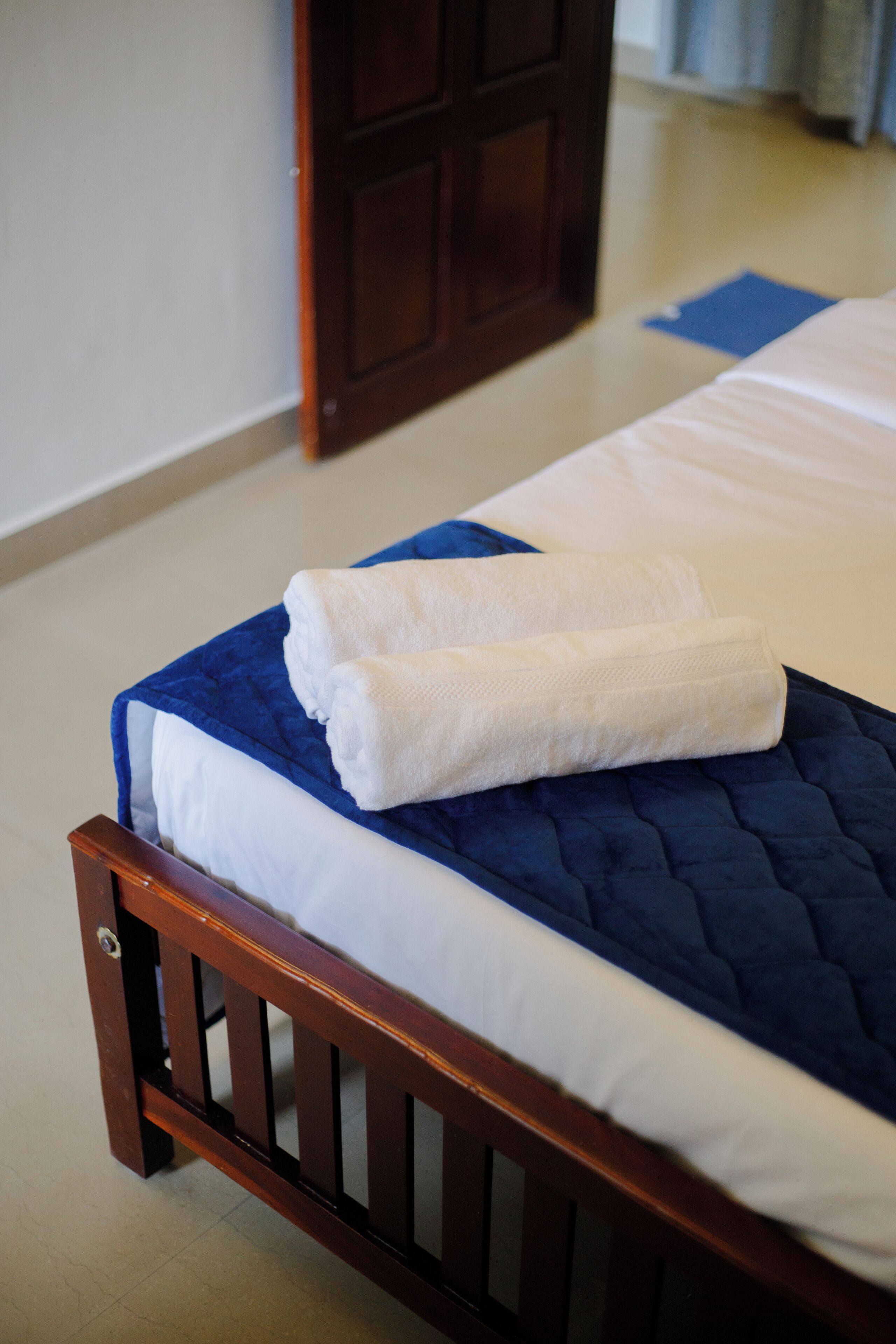 A neatly made bed with a blue quilt and white linens, a wooden headboard, and a blue mat on the floor.