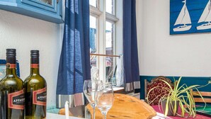 Ein Essbereich mit zwei Weinflaschen, zwei Gläsern, einem Holztisch, einem blauen Vorhang, einem Fenster mit Aussicht und einem Segelbootgemälde an der Wand.