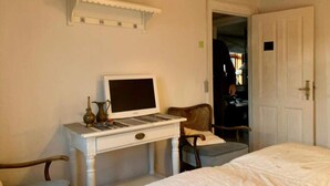 Ein Schlafzimmer mit einem Bett, einem Schreibtisch mit Computer, einem Sessel und einer Tür zu einem anderen Raum.