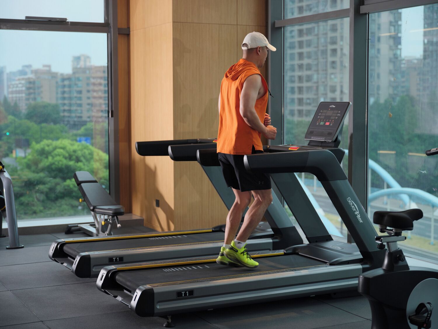 A person running on a treadmill with a digital display, in a gym with large windows and a view of buildings and trees.