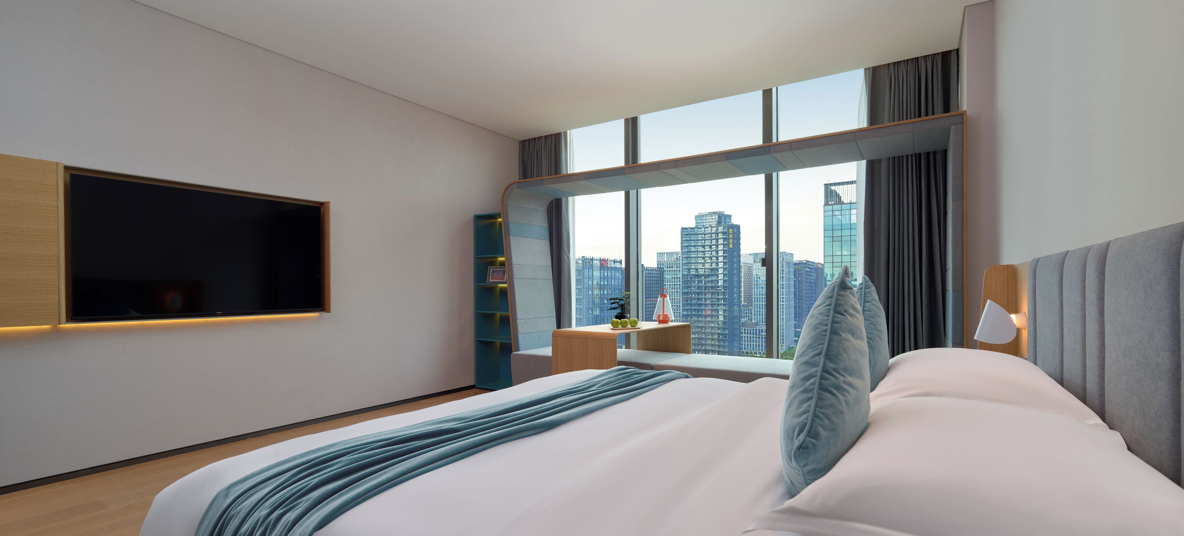 Ein modernes Hotelzimmer mit einem großen Bett, einem an der Wand montierten Fernseher, Blick auf die Stadt durch große Fenster und einer kleinen Sitzecke mit Tisch und Stühlen.