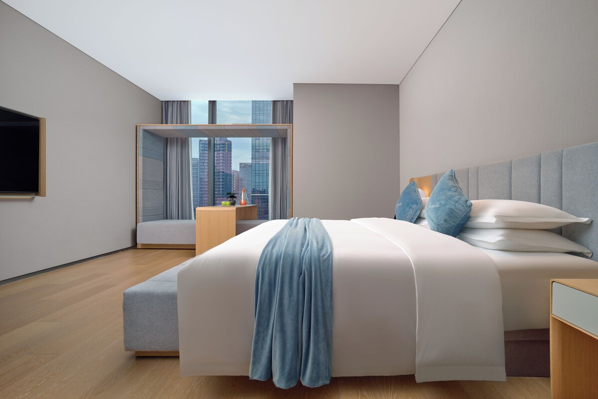 A modern bedroom with a large bed, a bedside table, a TV mounted on the wall, and a view of the city through large windows.