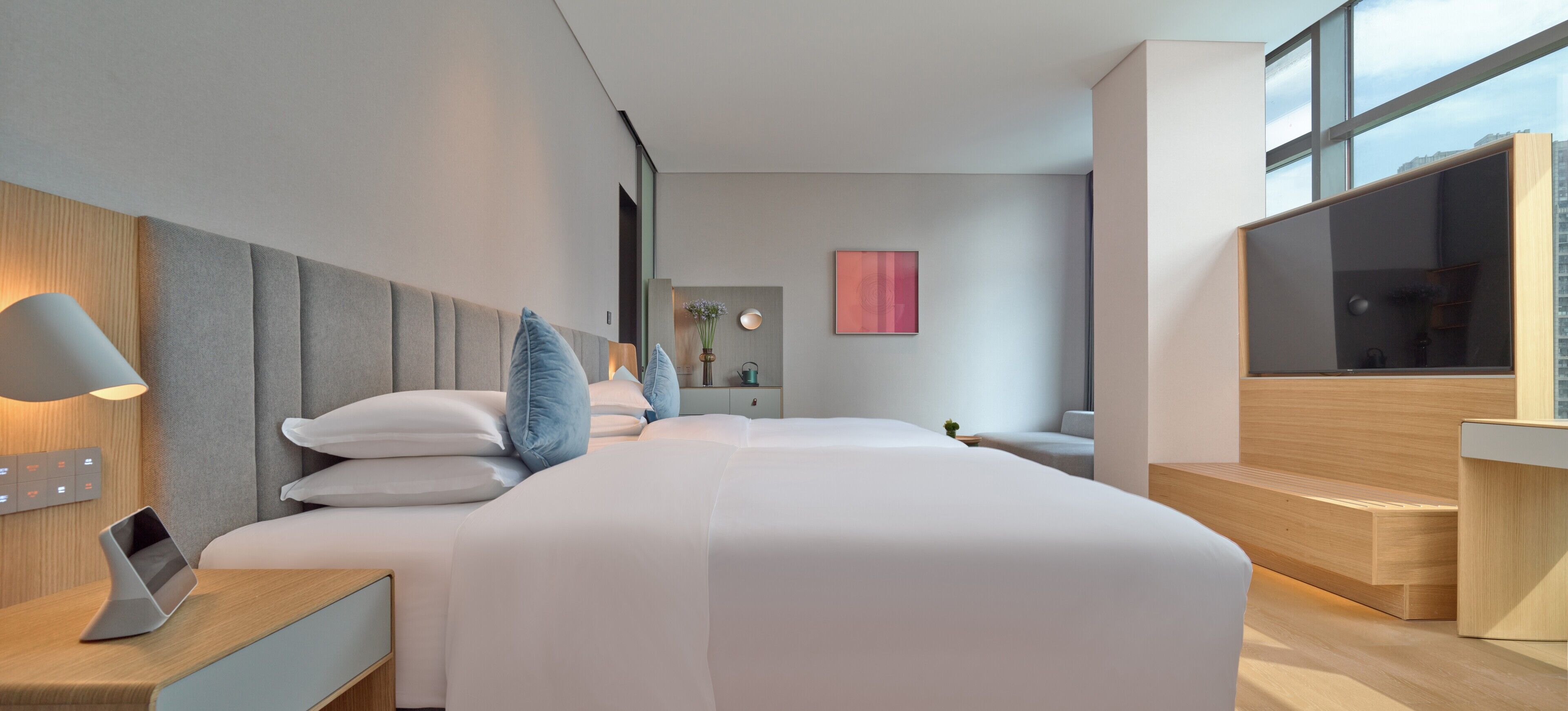 Quarto de hotel moderno com cama, mesa de cabeceira, abajur e uma janela ampla.
