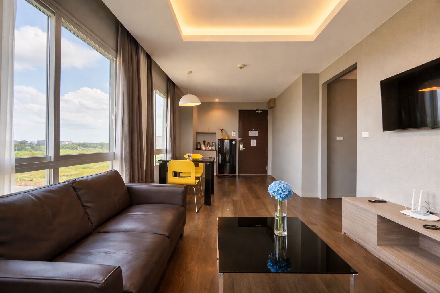 Une chambre d’hôtel moderne avec un canapé en cuir, un coin repas avec des chaises jaunes, une cuisine, un téléviseur à écran plat et une grande fenêtre donnant sur un paysage agréable.