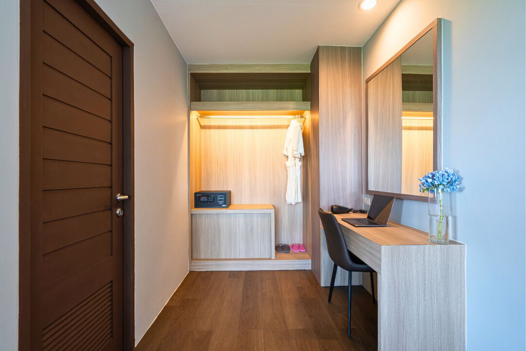 A hotel room with a wooden door, a desk with a laptop, a mirror, and a small plant.