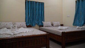 A room with two wooden beds, each with a patterned bedspread and pillows. Blue curtains are drawn on the windows. - Sonajhuri Bagan Bari (Bolpur)