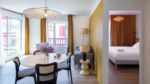 Habitación de hotel con cama, zona de comedor con mesa y sillas, lámpara y vista al exterior.