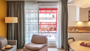 Una sala moderna con un sofá, una silla, una mesa de comedor con un libro y pasteles, y vistas a un balcón con barandillas rojas.