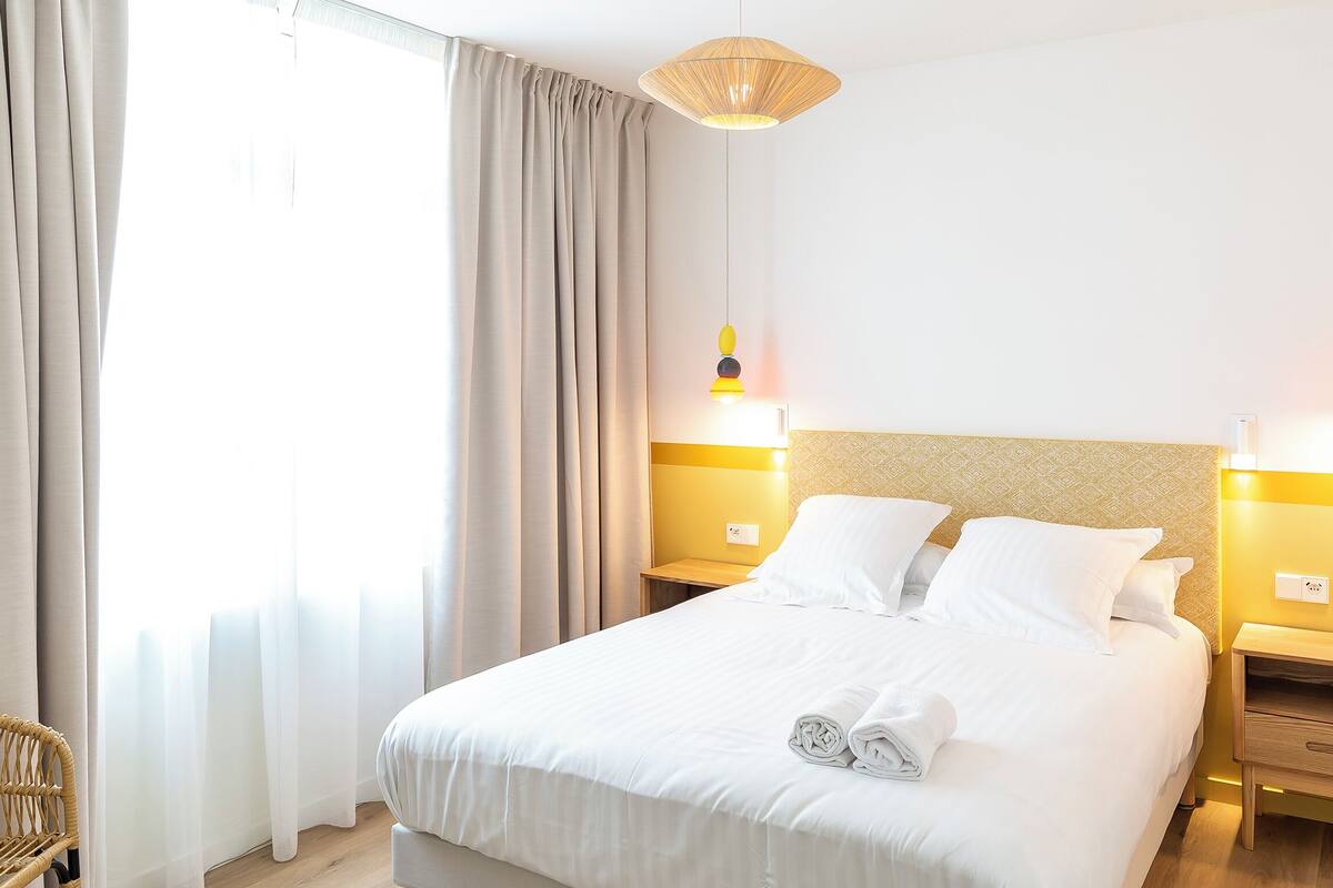 A neatly made bed with white linens, two bedside tables, a hanging light fixture, and a window with curtains.