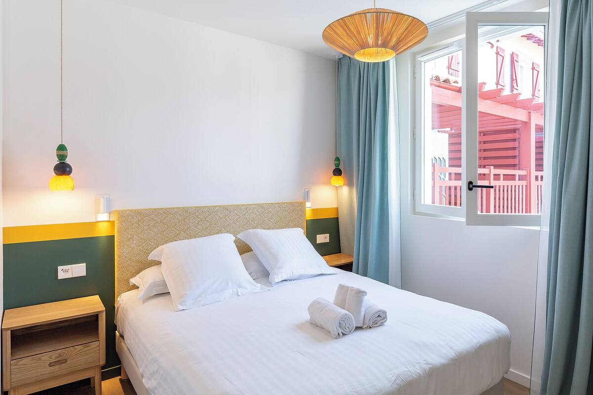 A neatly made bed with white linens and pillows, a bedside table with a lamp, and a view of a red building through a window with blue curtains.