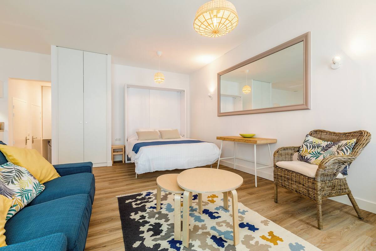 A modern bedroom with a bed, a blue sofa, a wicker chair, a small table, and a large mirror.