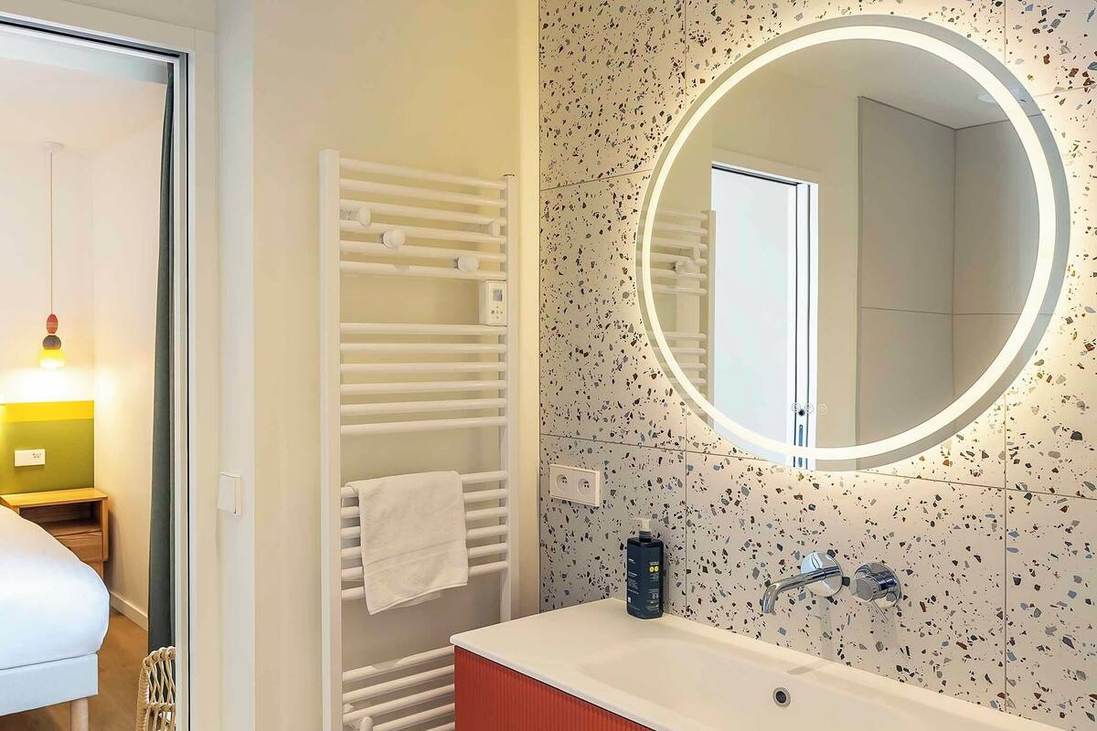 A hotel room with a bed, bedside table, lamp, towel rack, and a bathroom with a sink, mirror, and decorative wall.