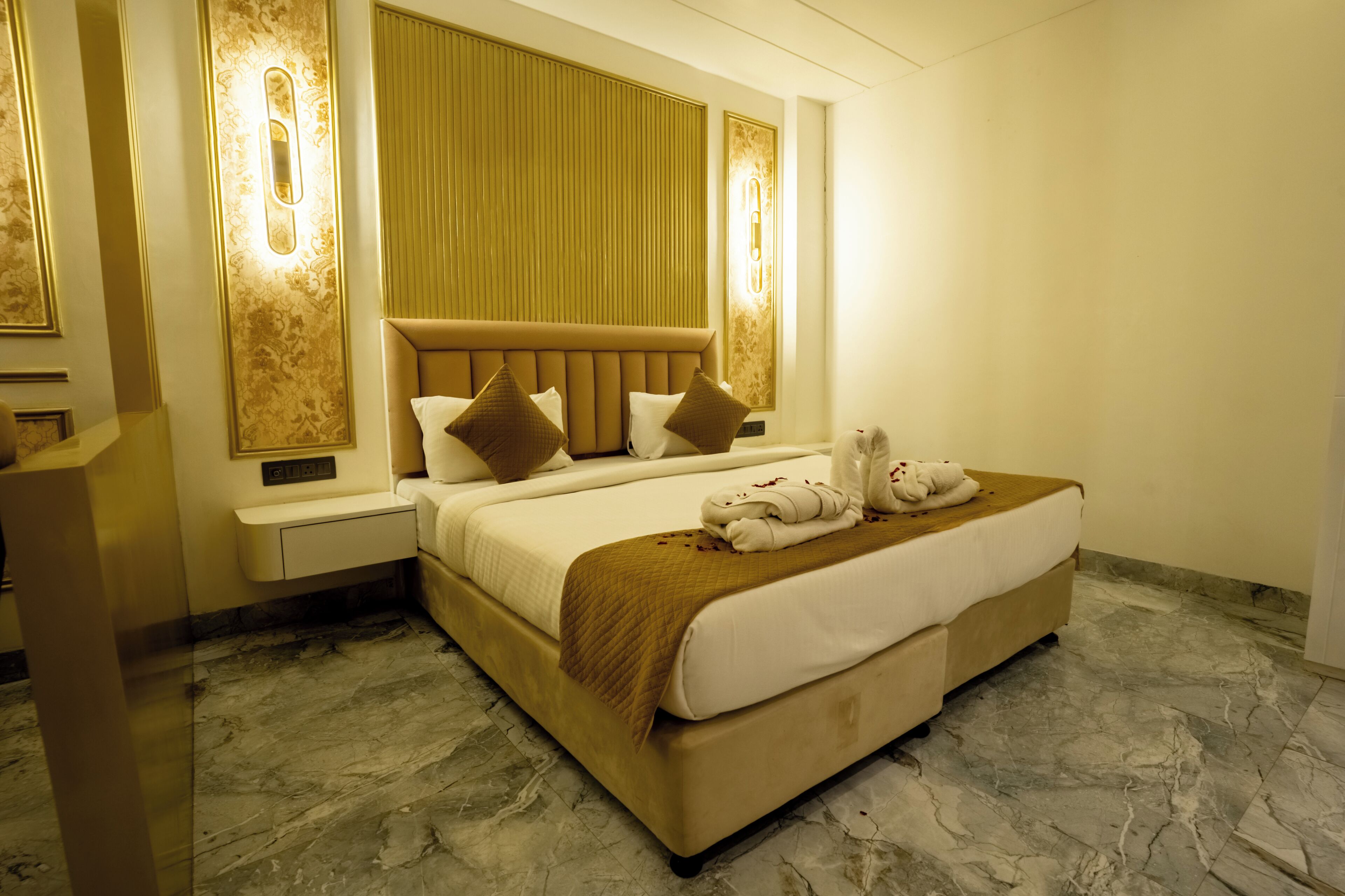 A hotel room with a large bed, a nightstand, a mirror, and a wall-mounted light fixture.