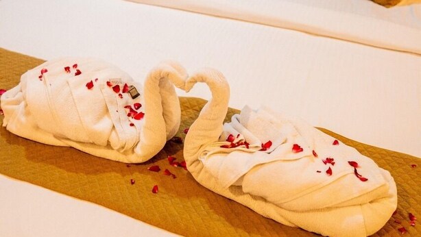 Un lit parfaitement fait, agrémenté d’une serviette pliée en forme de cygne et de pétales de rose disposés sur un plateau en bois.