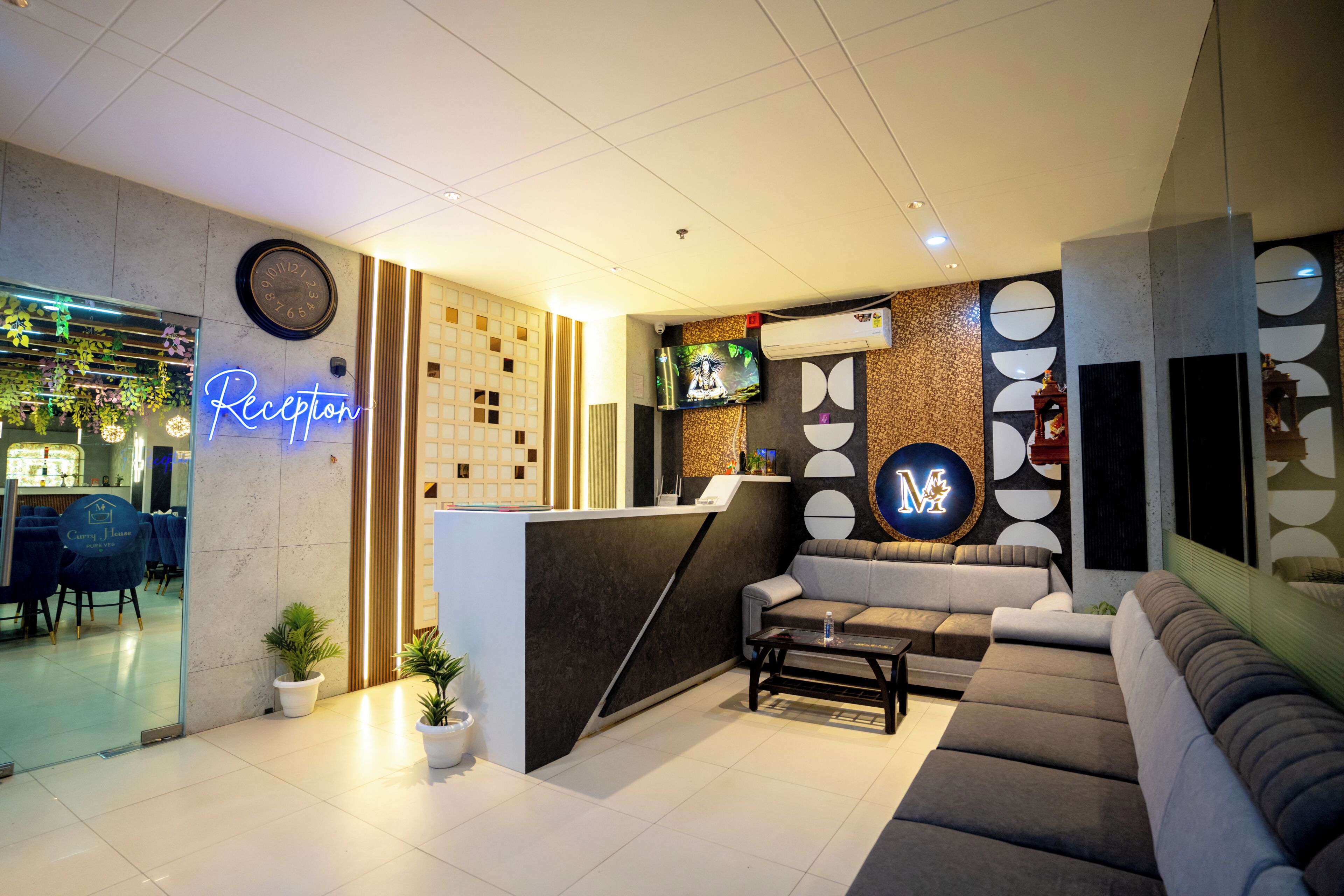 A reception area with a neon sign, a clock, a TV, a reception desk, sofas, and potted plants.