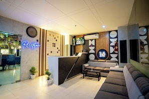 Ein Empfangsbereich mit einer Neonreklame, einer Uhr, einem Fernseher, einer Rezeption, Sofas und Topfpflanzen.