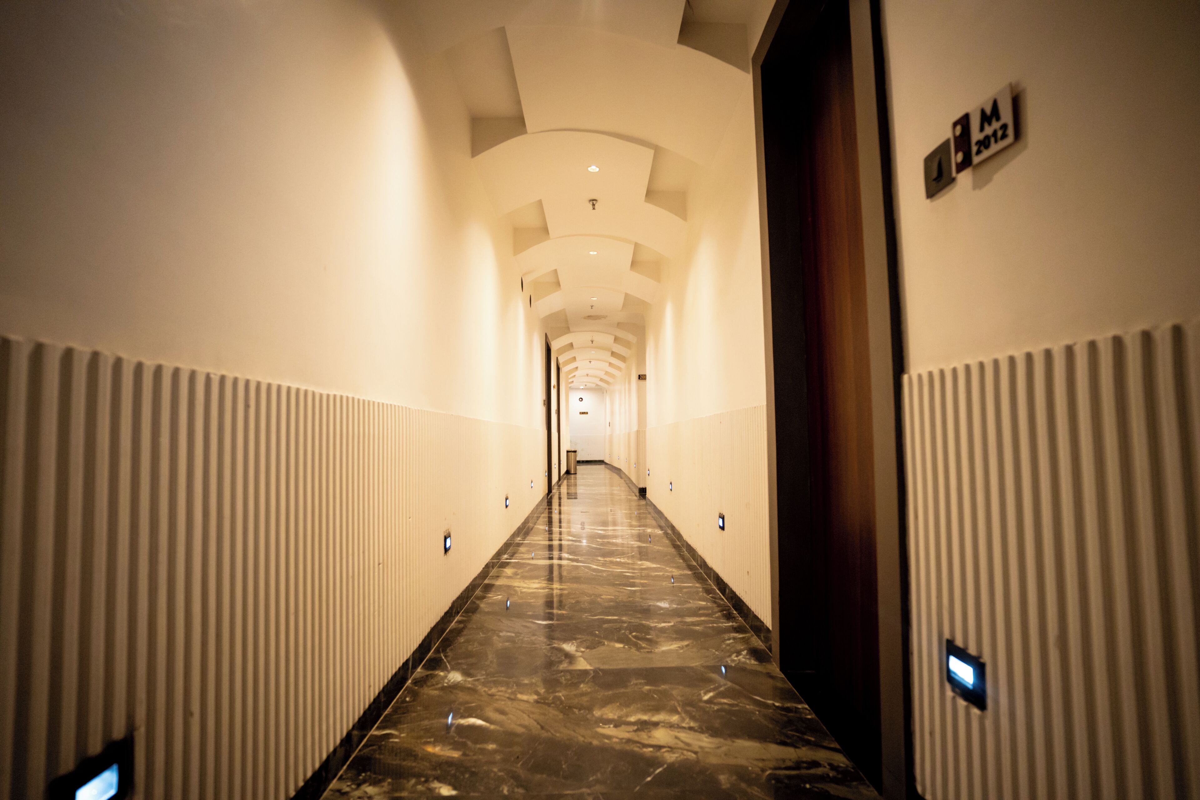 Um corredor com piso de mármore, paredes brancas e uma porta de madeira à direita.