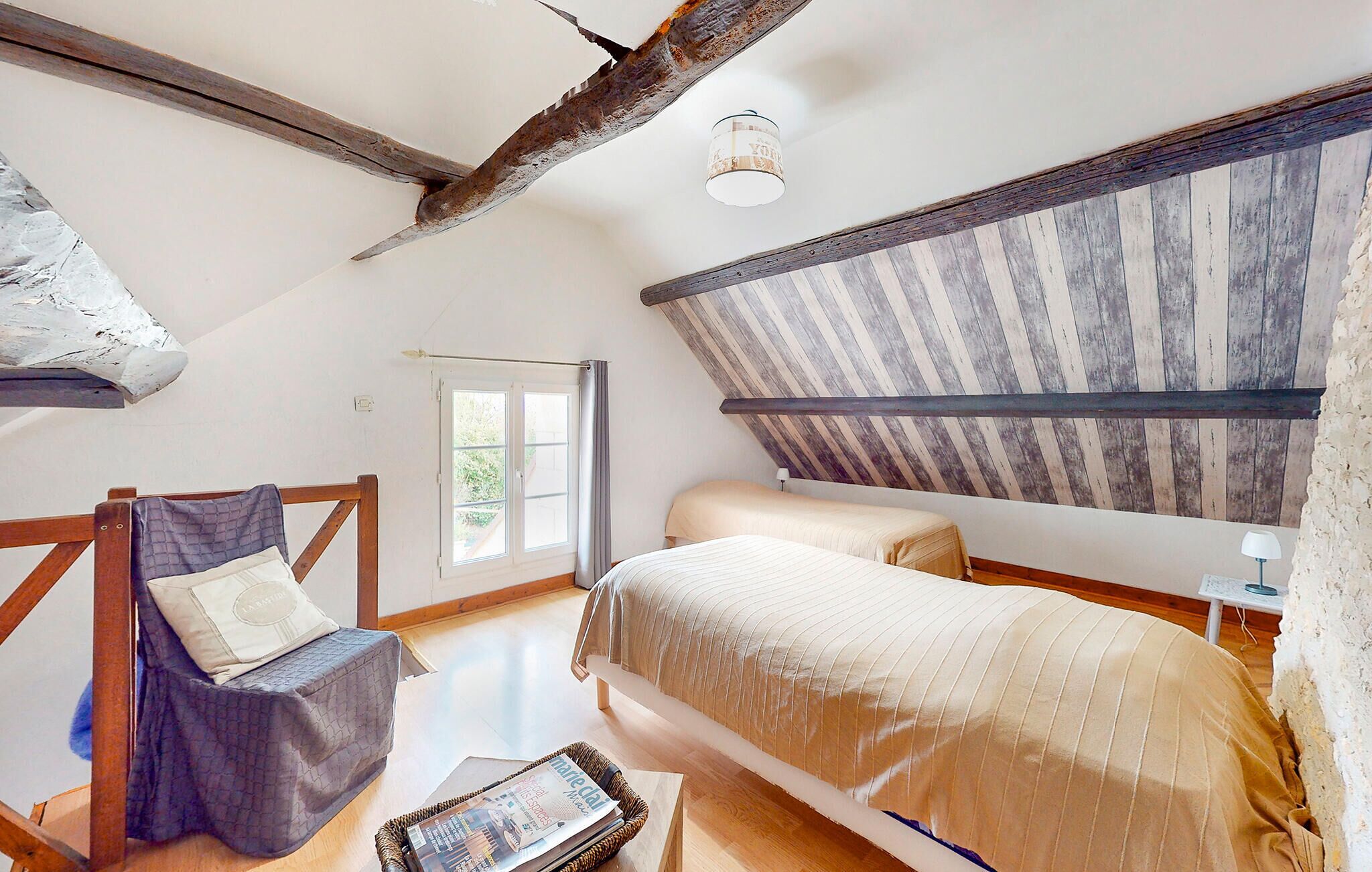 A bedroom with a sloped ceiling, a bed, a chair, a window, and a small table with a magazine.