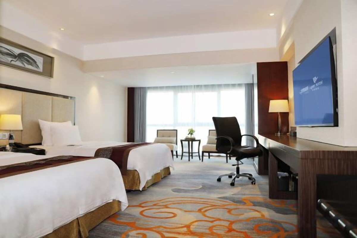 A hotel room with two beds, a desk, a chair, a television, and a large window.