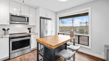 Une cuisine moderne avec des meubles blancs, des appareils en acier inoxydable, un îlot central en bois et une grande fenêtre donnant sur la ville.