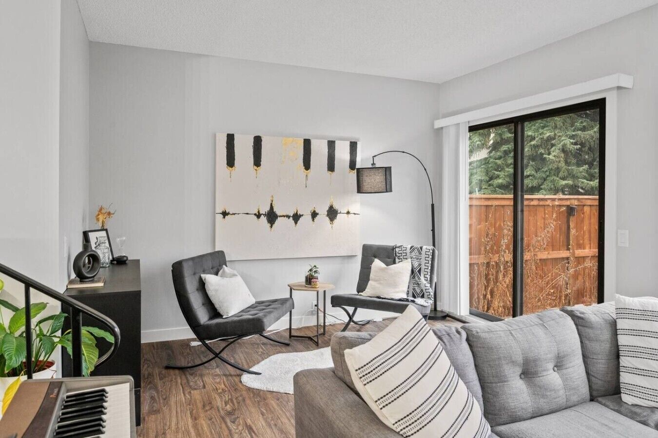 Un salon moderne avec un canapé gris, des fauteuils noirs, une petite table d’appoint et une grande fenêtre donnant sur une terrasse en bois.