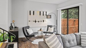 A modern living room with a grey sofa, black armchairs, a small side table, and a large window with a view of a wooden deck.
