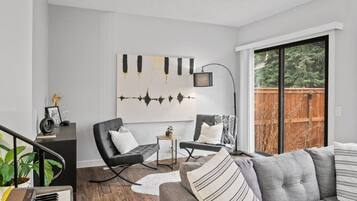 Un salon moderne avec un canapé gris, des fauteuils noirs, une petite table d’appoint et une grande fenêtre donnant sur une terrasse en bois.