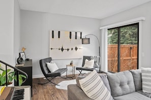 Uma sala de estar moderna com um sofá cinzento, poltronas pretas, uma mesa de apoio pequena e uma janela grande com vista para um deck de madeira.