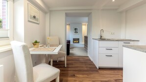 A modern kitchen with white cabinetry, a dining area with a table and chairs, and a fireplace in the background. - Sylt Rooms 10 - Shelter Island - Westerland (Westerland)