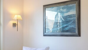 A room with a bed, white pillows, a wall-mounted lamp, and a framed painting of a sailboat on the sea. - Sylt Rooms 10 - Shelter Island - Westerland (Westerland)