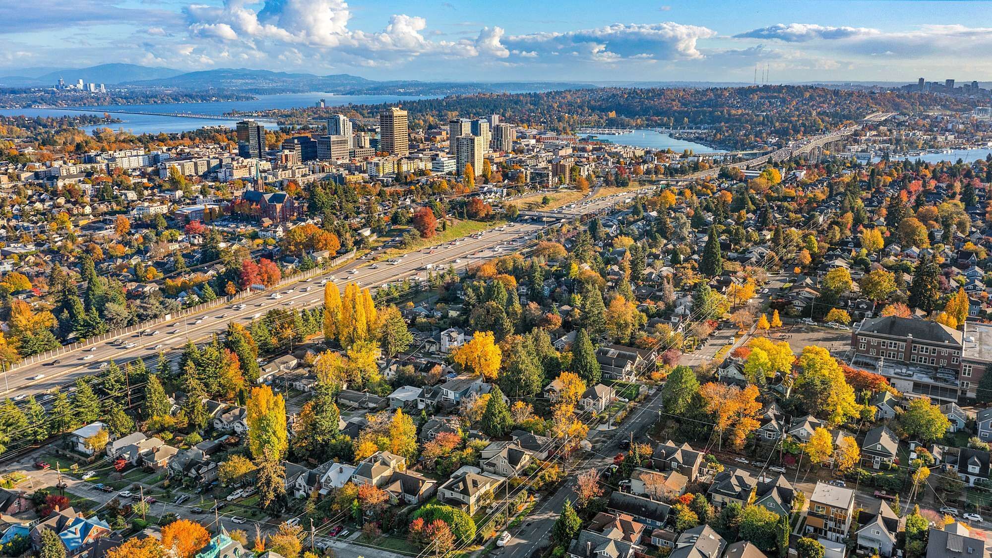 An aerial view of a city with dense residential areas, a major highway, and a large body of water in the background.