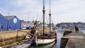 A harbor with a large sailing ship docked, a blue building with a logo, and a waterfront promenade.