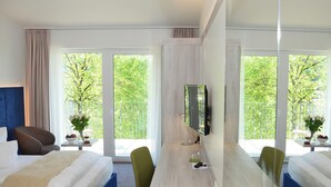 Ein Hotelzimmer mit Bett, Schreibtisch, Stuhl, Fernseher und großen Fenstern mit Blick auf Bäume.