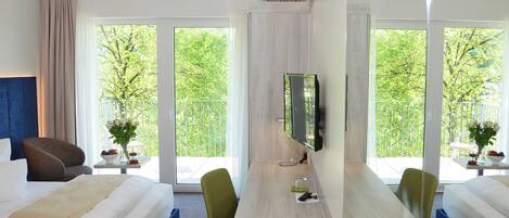Habitación de hotel con cama, escritorio, silla, televisor y ventanas amplias con vistas a árboles.
