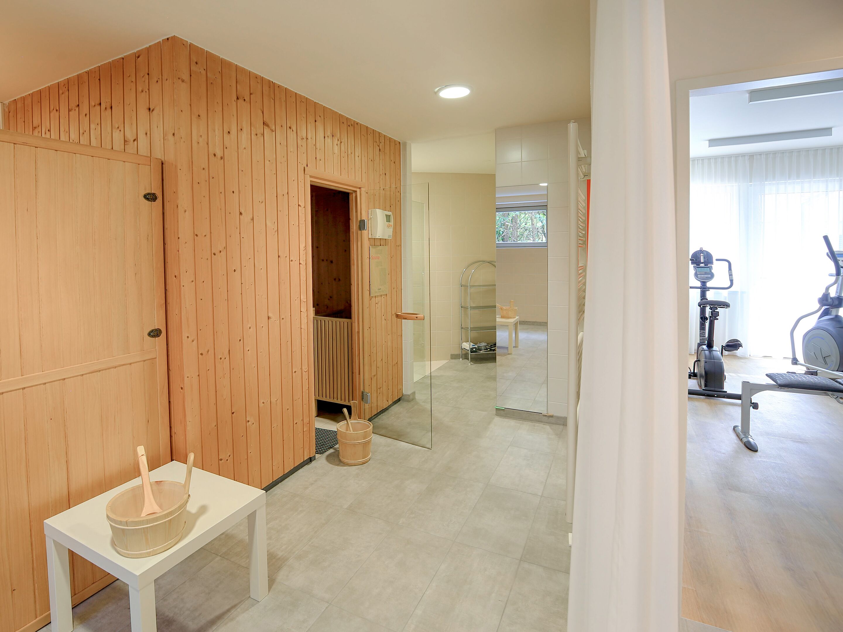 Una habitación con pared de madera, una mesita con un cubo de madera, una puerta de vidrio que da a un baño y equipo de ejercicio en la habitación contigua.