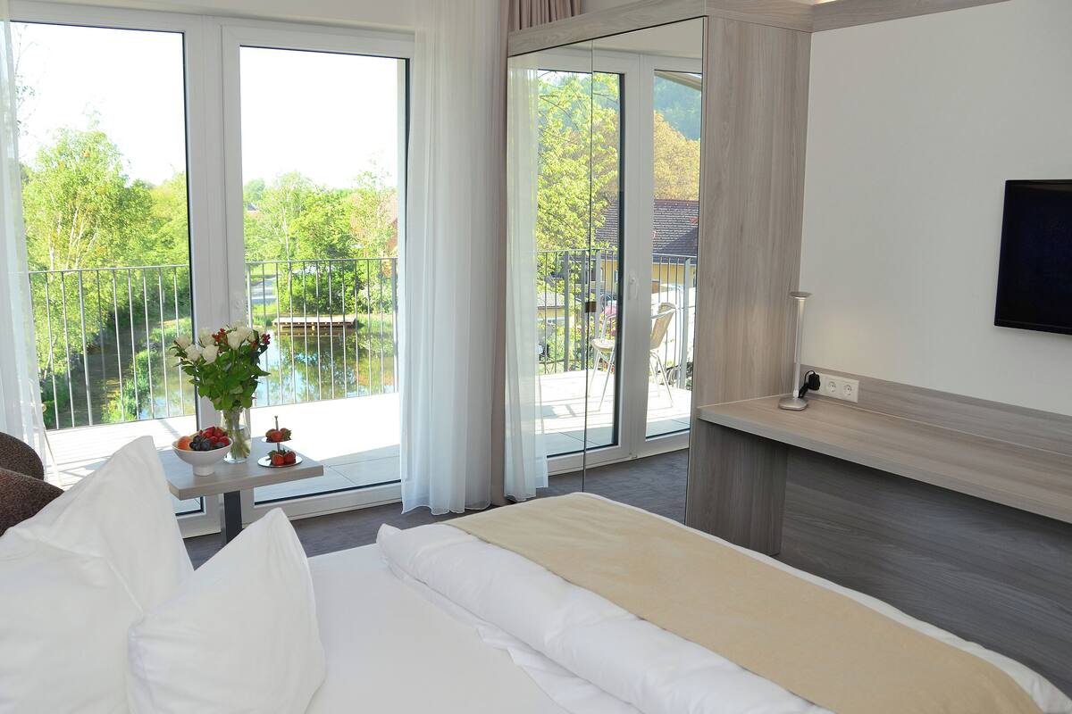 A hotel room with a large bed, a TV mounted on the wall, and a balcony with a view of greenery and a body of water.