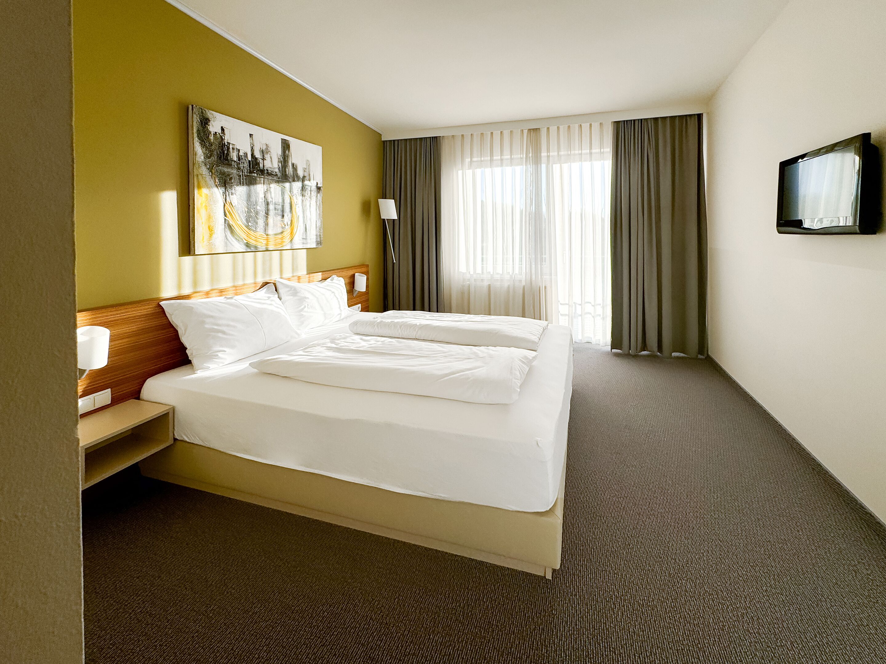 Une chambre d’hôtel avec un grand lit, un tableau au mur, un téléviseur fixé au mur et une fenêtre avec des rideaux.