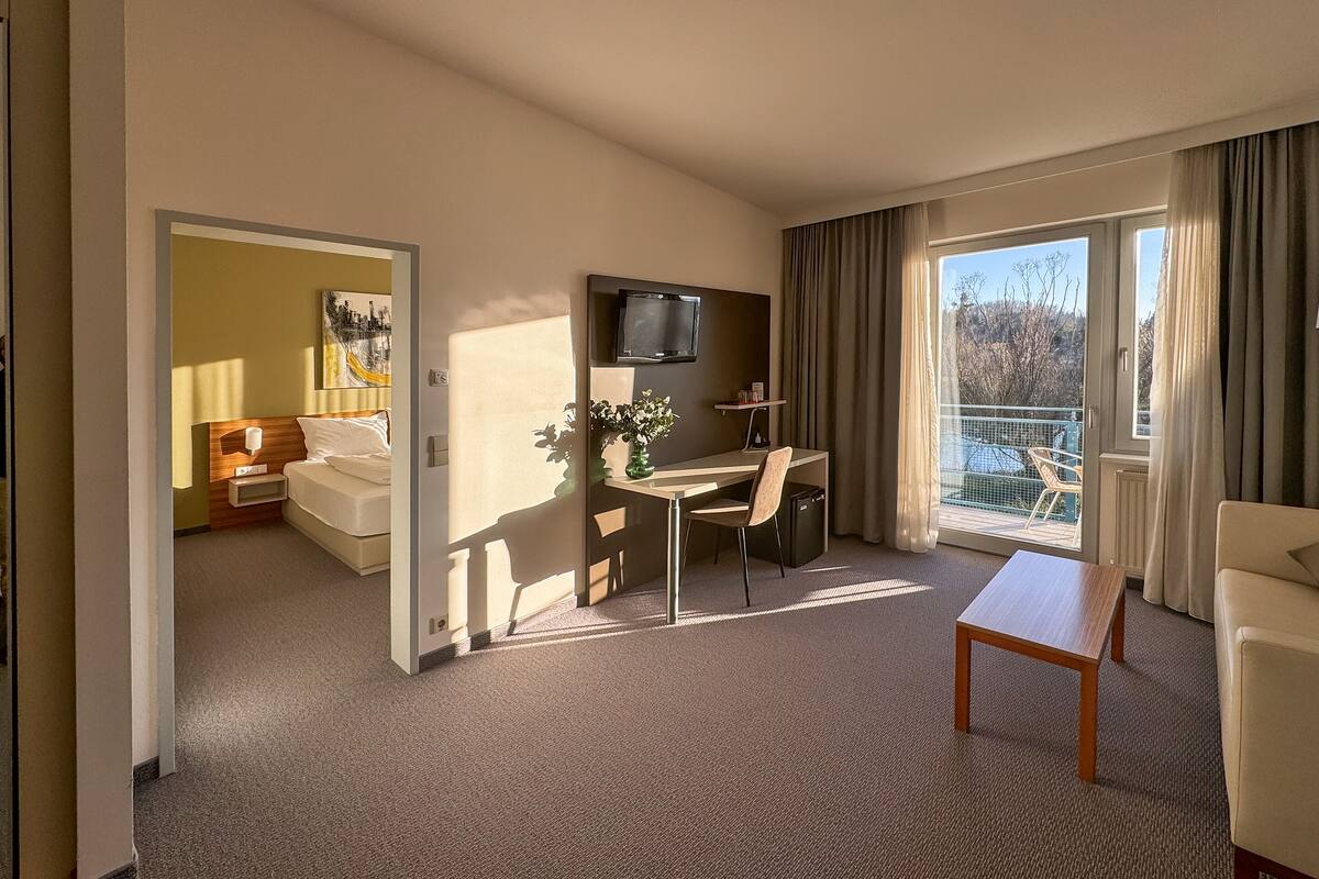 A hotel room with a bed, a desk with a chair, a TV, and a balcony with a view.