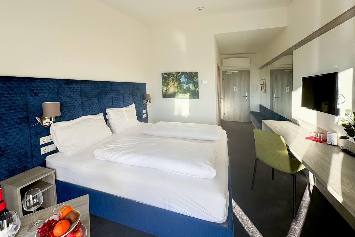 A hotel room with a large bed, a desk with a chair, a TV, and a small table with a fruit bowl and glasses.